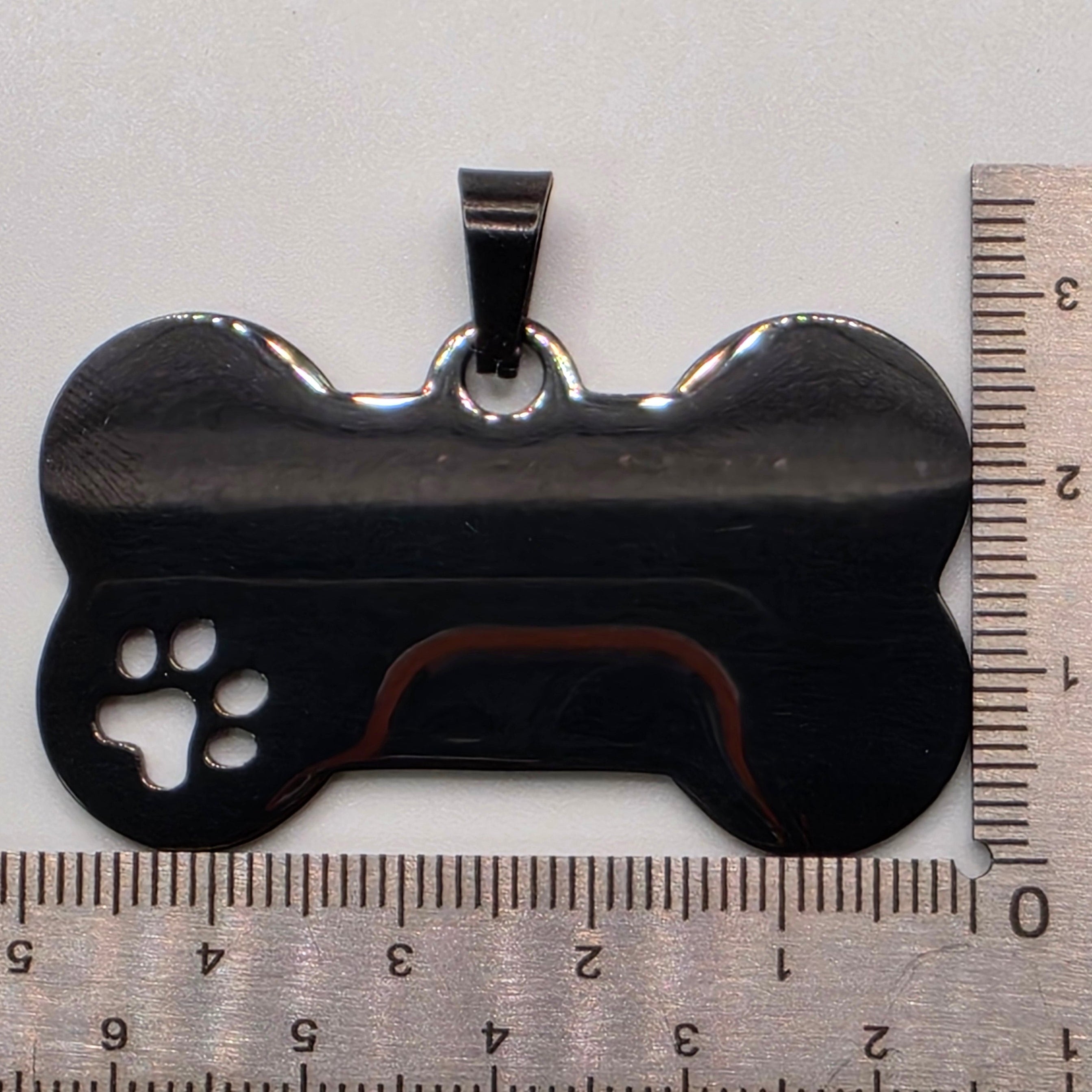 Black metal dog bone-shaped tool with paw print cutout next to a ruler on a white background
