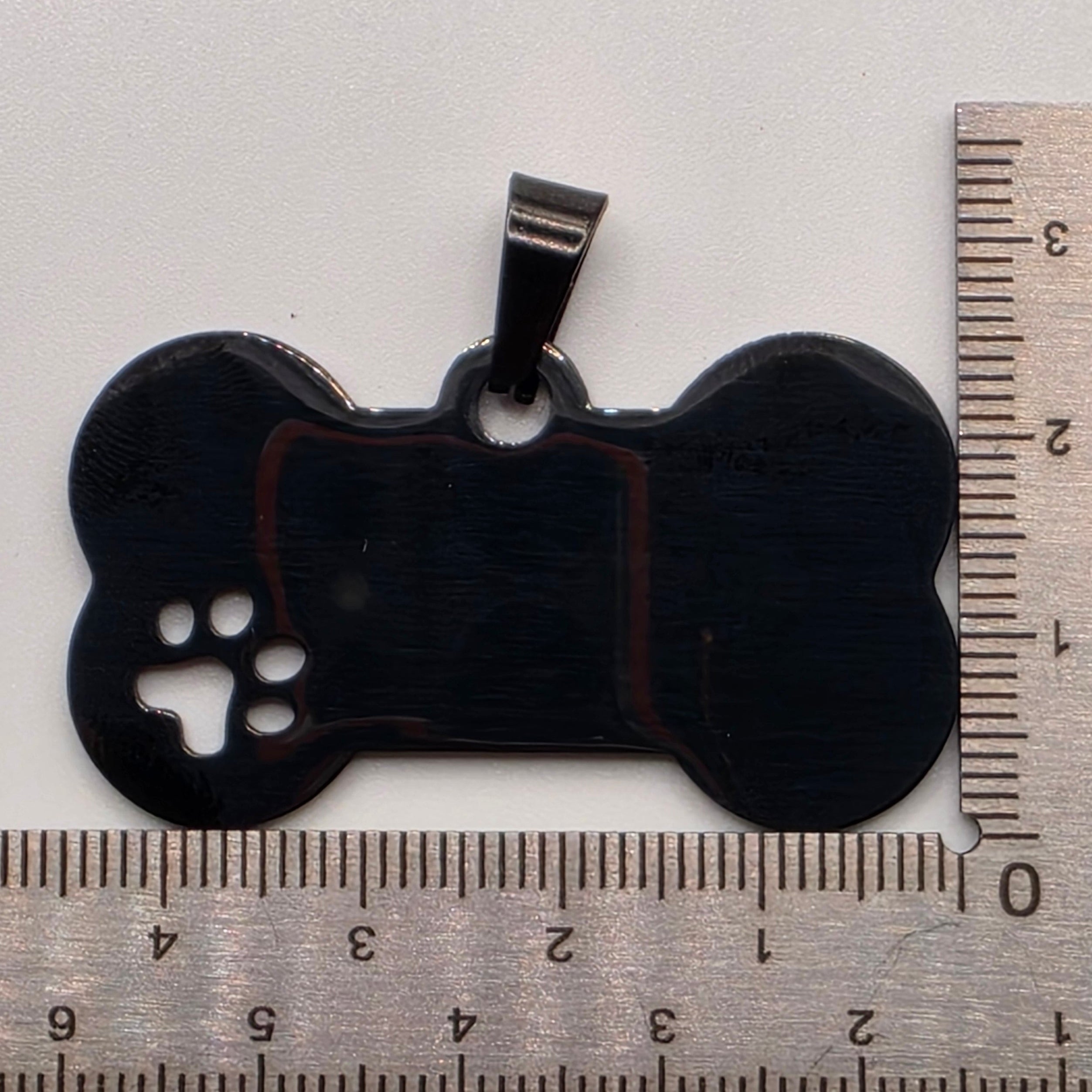 Black bone-shaped pet tag with a paw print cutout next to a ruler on a white background