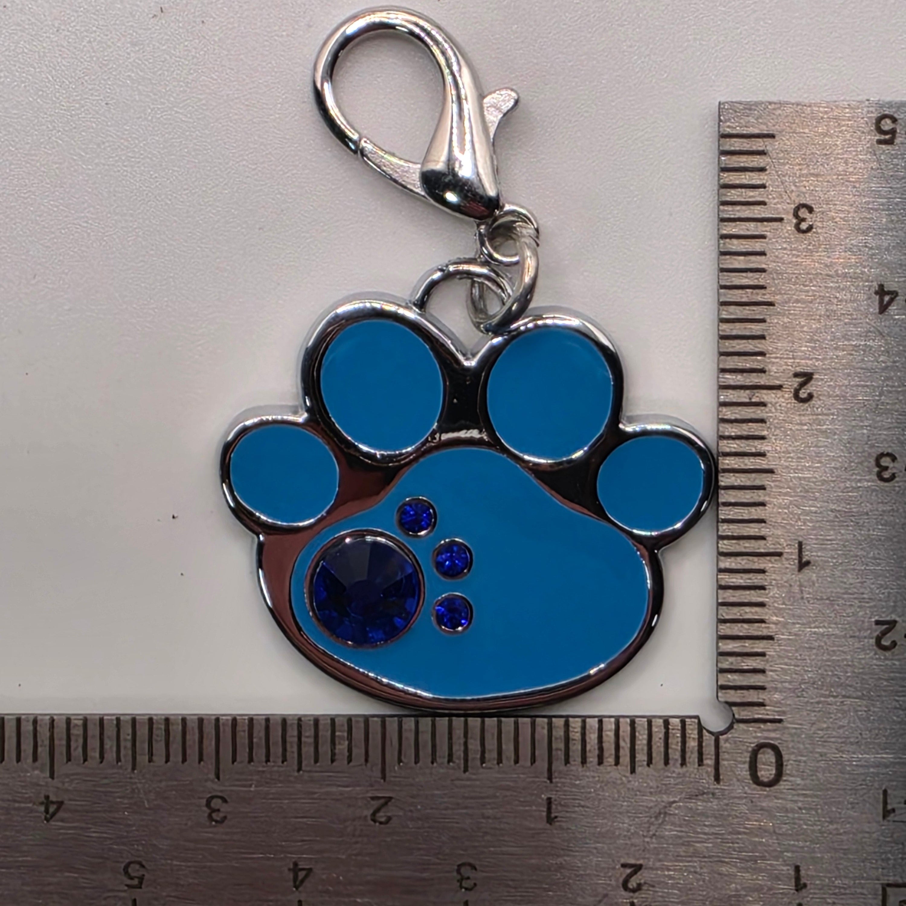 Blue paw print charm with a clasp on a white background, measured against a ruler.
