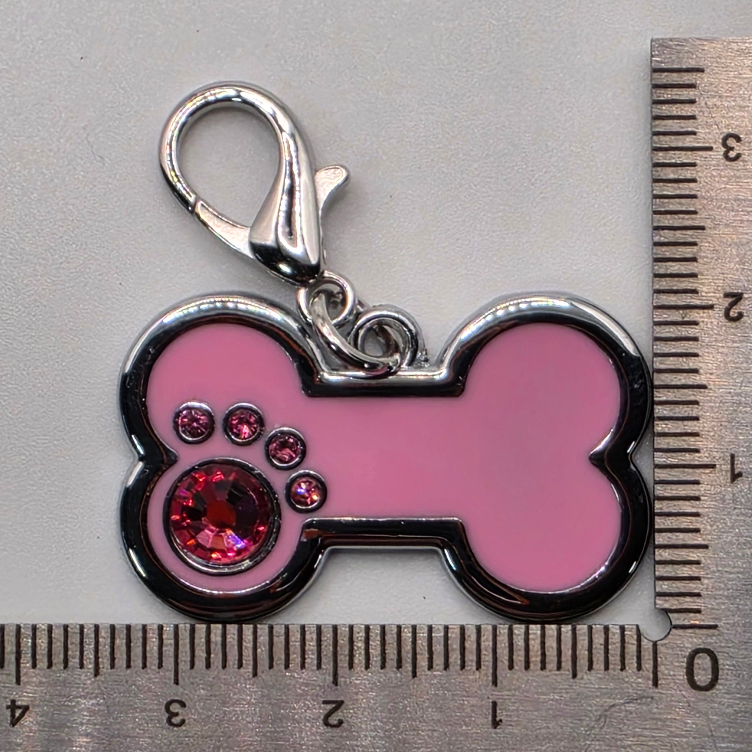 Pink bone-shaped keychain with a silver clasp and red gemstone, measured against a ruler on a gray background.