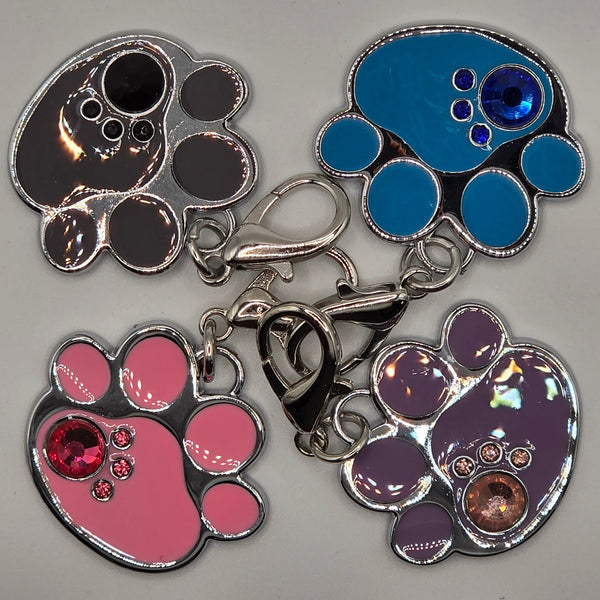 Four paw-shaped charms in blue, purple, black, and pink with embedded stones on a gray background.