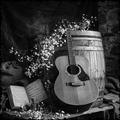 Acoustic guitar leaning against a wooden barrel with flowers and books on a stone surface.
