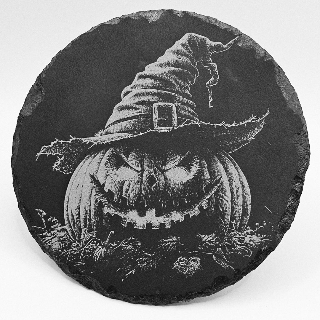 Black slate coaster with a carved pumpkin and witch's hat design on a white background